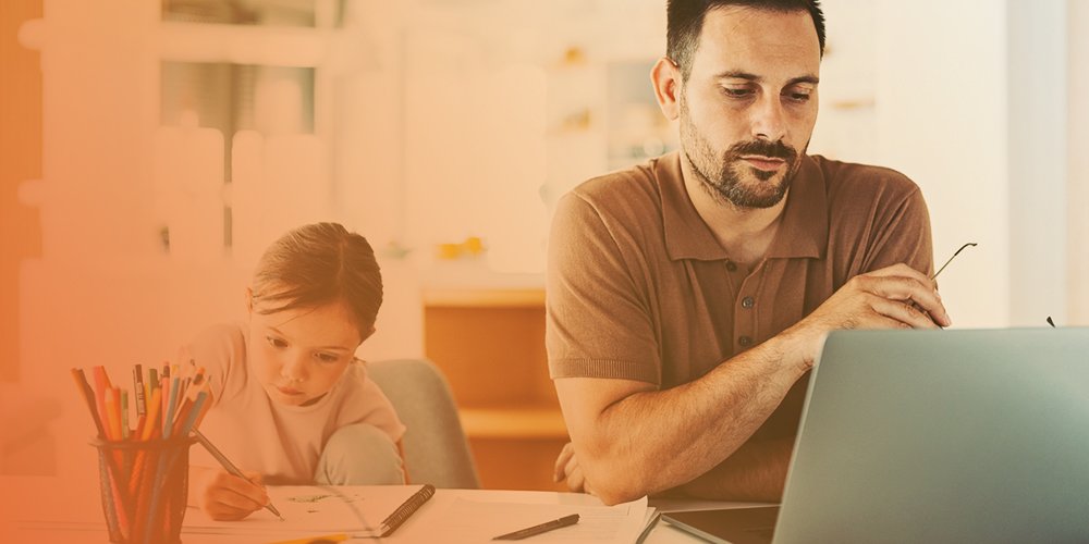 Man on laptop working while child colours on pad of paper next to him on table