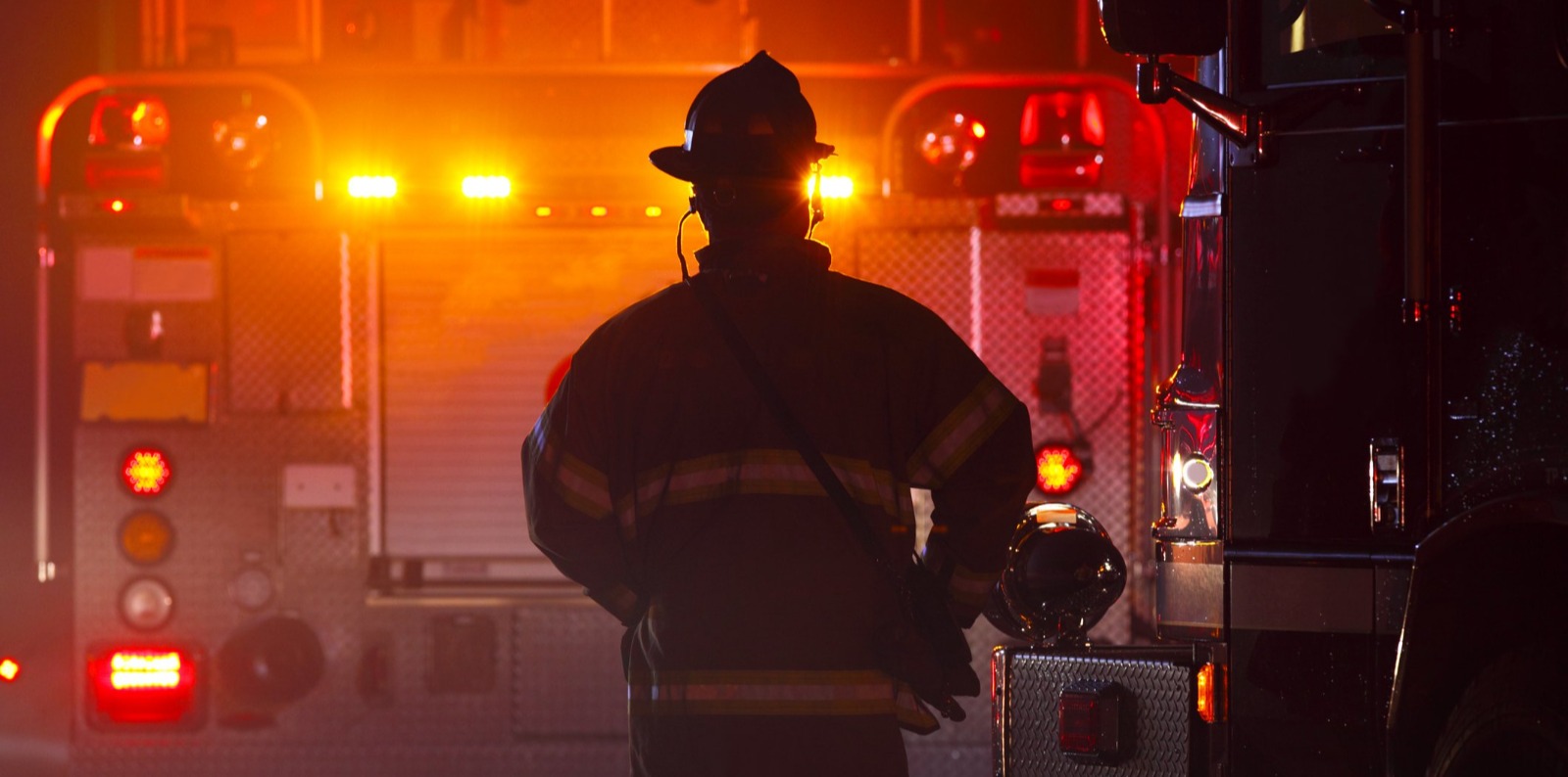 Firefighter silhouetted against a fire truck with flashing lights at an emergency scene